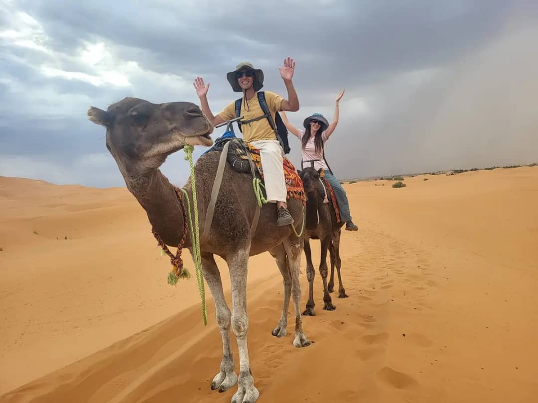Couple waving from their camels on the Merzouga dunes under a stormy sky