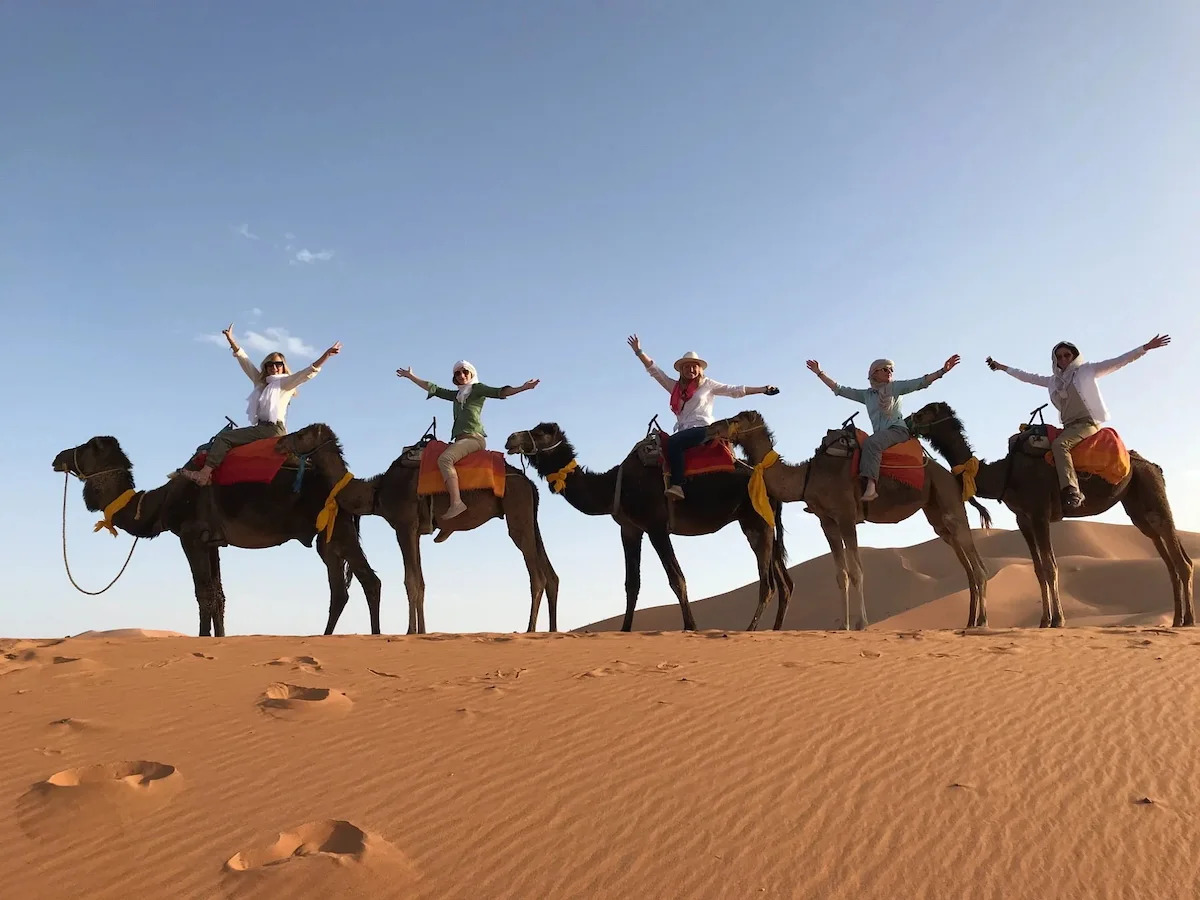 Happy client riding a camel at sunset in the Sahara Desert, silhouetted against the golden Erg Chebbi dunes during our Morocco camel tour